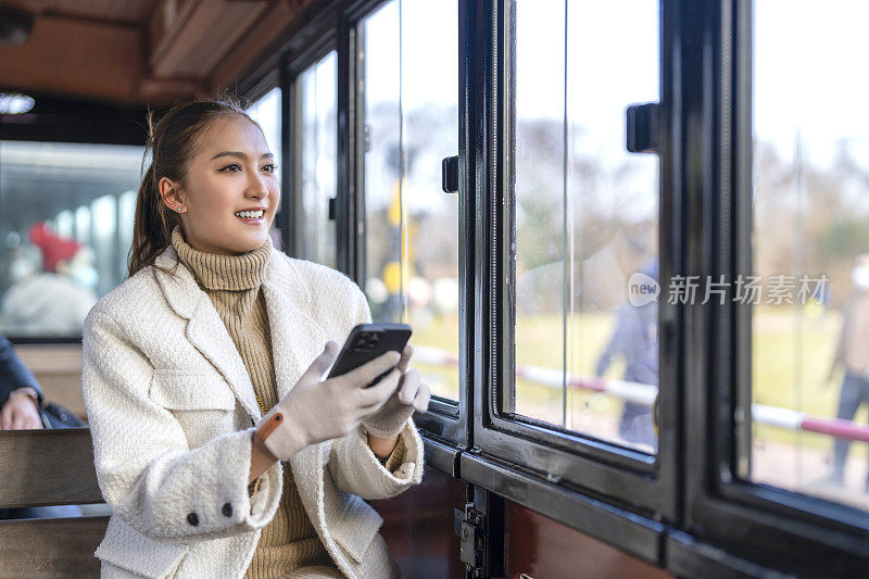 幸福的亚洲女人独自旅行坐在穿梭轨道列车上看外面的花园公园景色，亚洲女人在韩国济州岛旅行时用智能手机拍照