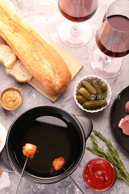 火锅锅、叉子上放着炸好的肉片、红酒酒杯等制品放在灰色的有质感的桌子上，平铺着