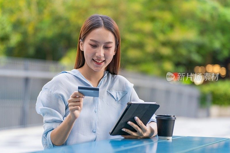 一名亚洲妇女在户外公园用数字平板电脑进行数字银行信用卡交易，桌上放着一杯咖啡