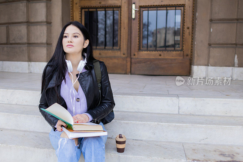 年轻漂亮的女大学生背着双肩包坐在大学楼梯上。她戴着耳机，穿着皮夹克，等待着大学课程的开始。