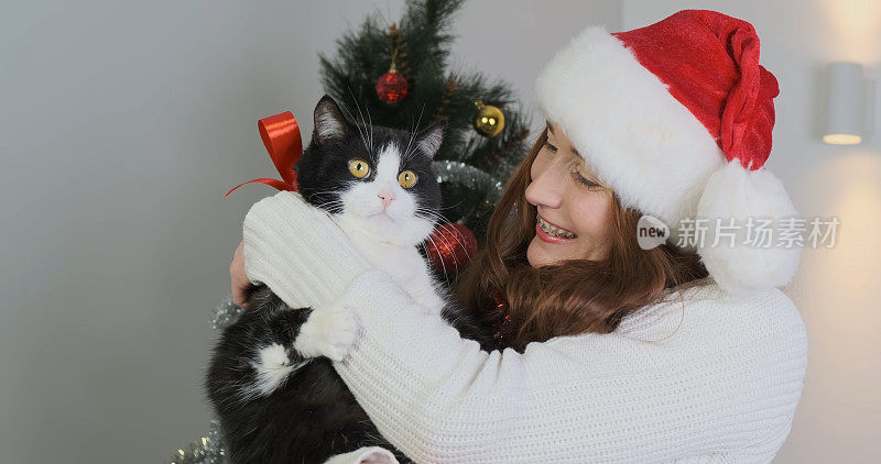 一位戴着新年帽的年轻女子，在一棵装饰好的圣诞树前，抱着一只有天赋的猫。这只黑白相间的猫系着一条红色的礼物丝带。