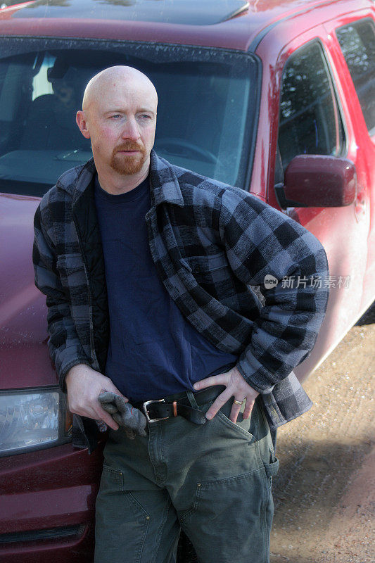 一个穿着格子衬衫的秃顶男人倚靠在一辆肮脏的红色卡车上