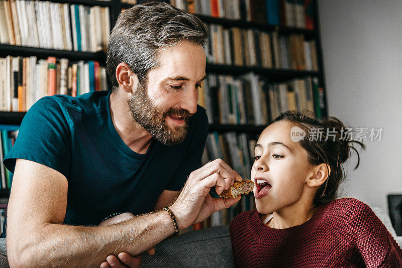 长胡子的父亲在沙发上喂女儿吃三明治