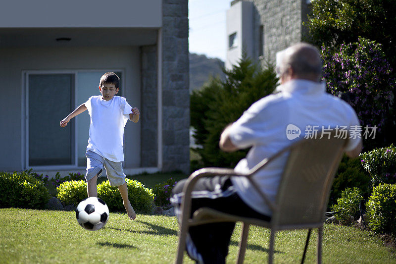 爷爷坐在椅子上，他的孙子在踢足球