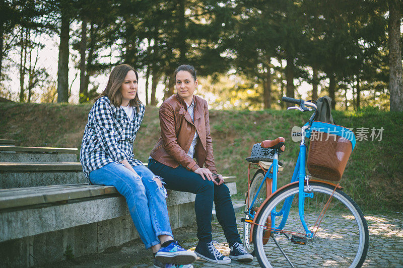 两个可爱的快乐的微笑美丽的年轻女自行车朋友穿着休闲衣服坐在公园长椅上在夏天的一天，有美好的时间在一起，