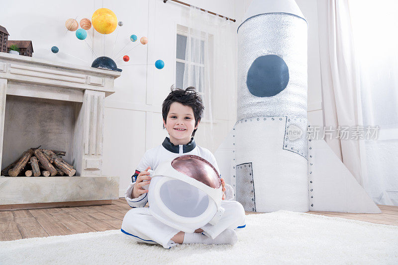 穿着宇航员服装的男孩拿着头盔，后面是玩具火箭