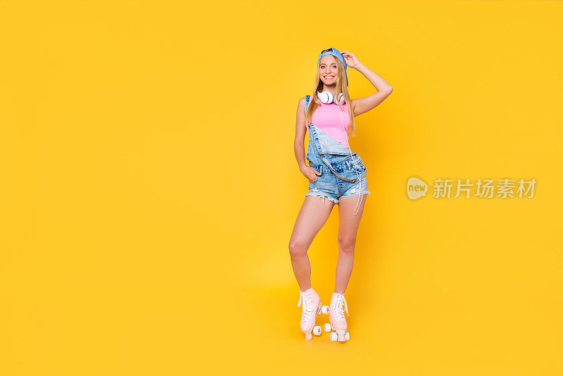 苗条漂亮的女孩的肖像在牛仔整体棒球帽戴在脖子上溜冰鞋手在短裤口袋看在黄色背景孤立的相机