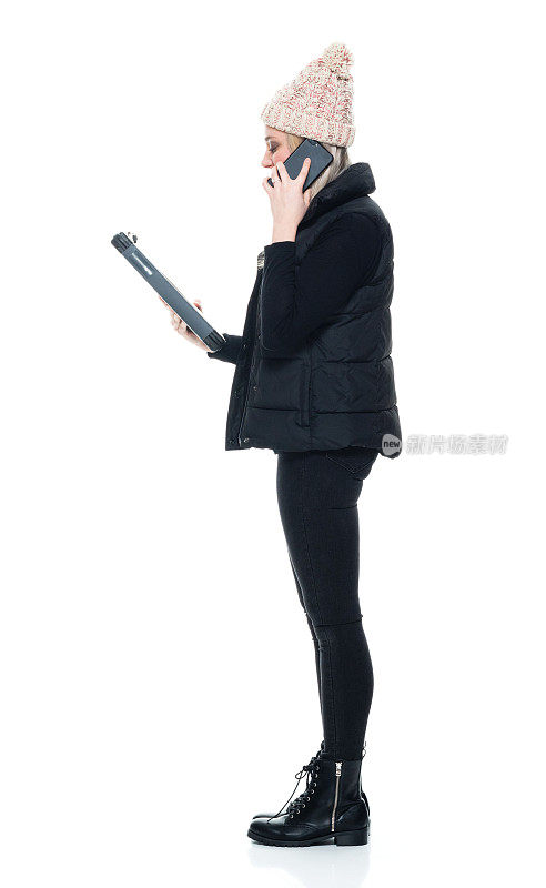 白种年轻女性无袖站在白色背景前，穿着靴子，手持文件，使用手机