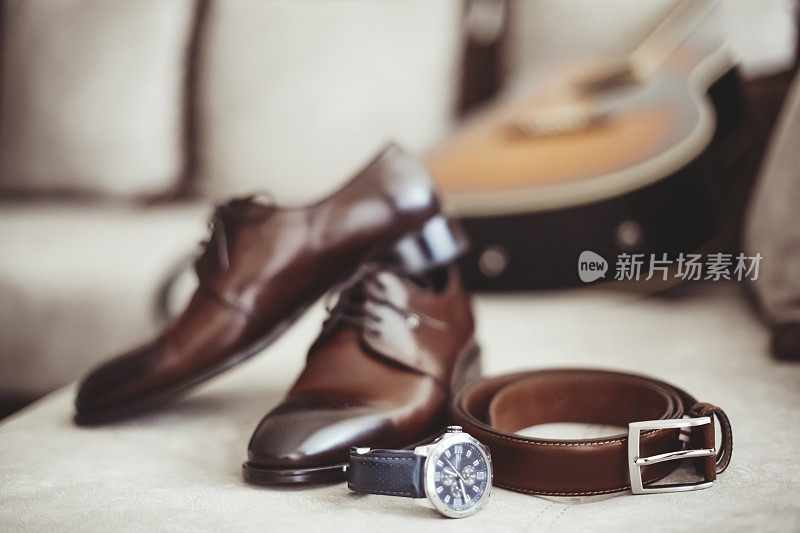 男性配饰包括相配的棕色皮带和鞋子，还有腕表。背景是一把吉他