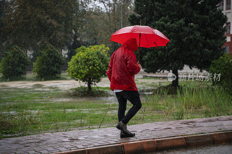 成熟的男人穿着红雨衣，撑着红伞走在雨中的城市街道上