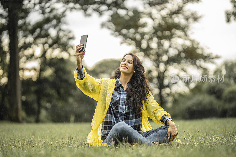 美丽的年轻女子在黄色雨衣与她的手机自拍在自然