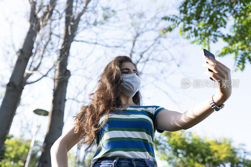 在新冠肺炎大流行期间，妇女在户外佩戴可重复使用的手工口罩。一个女孩在空旷的公园里用手机自拍。保持安全。