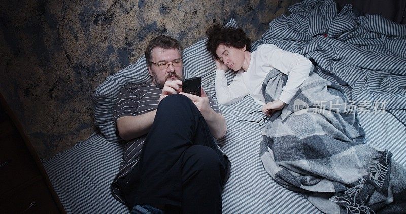 成熟的妻子试图睡觉，而丈夫在床上使用她的智能手机