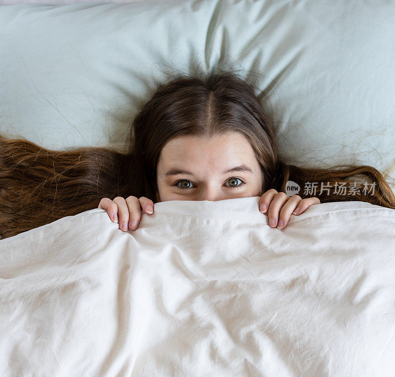 一个顽皮害羞的金发年轻女人，躲在被子下，躺在床上的羽绒被