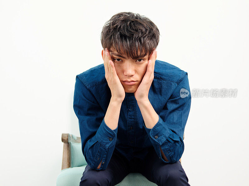 英俊的中国年轻男子的肖像与卷曲的黑色头发，蓝色衬衫坐在扶手椅和摆姿势的白墙背景。手放在脸上，看起来很焦虑，前视摄影棚拍摄