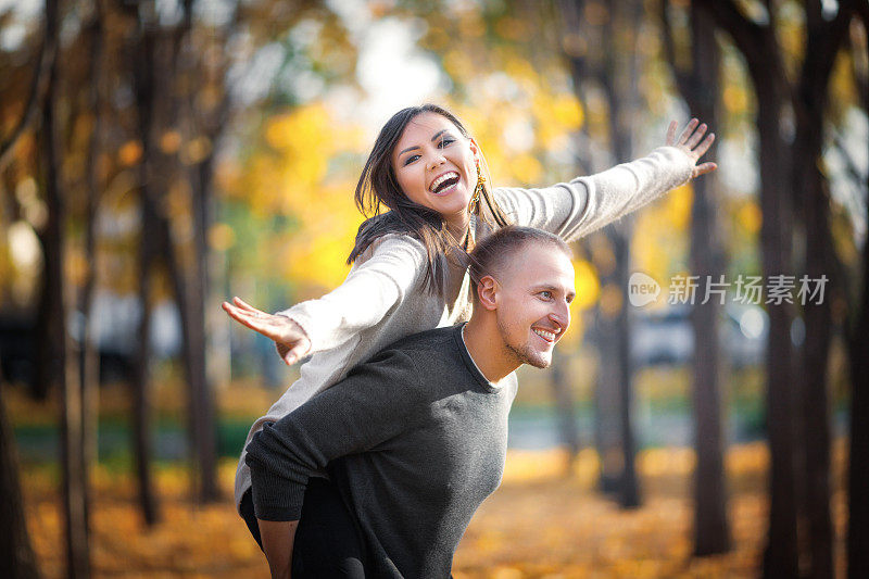 倾心的年轻夫妇在秋天的户外嬉戏。一个男人让一个女人躺在他的背上，她描绘了一架飞机
