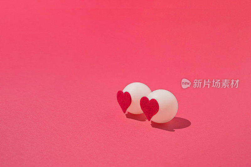 两个白色的乒乓球，粉红色的背景上有红心。最小限度的爱或情人的概念。
