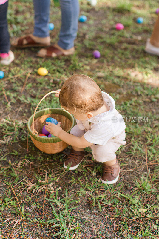 一个可爱的古巴裔美国15个月大的男婴，浅棕色头发和棕色眼睛，穿着棕色兔子和胡萝卜工作服和棕色鞋子，手拿复活节彩蛋篮，享受他的第一次复活节彩蛋寻找庆祝复活节假期