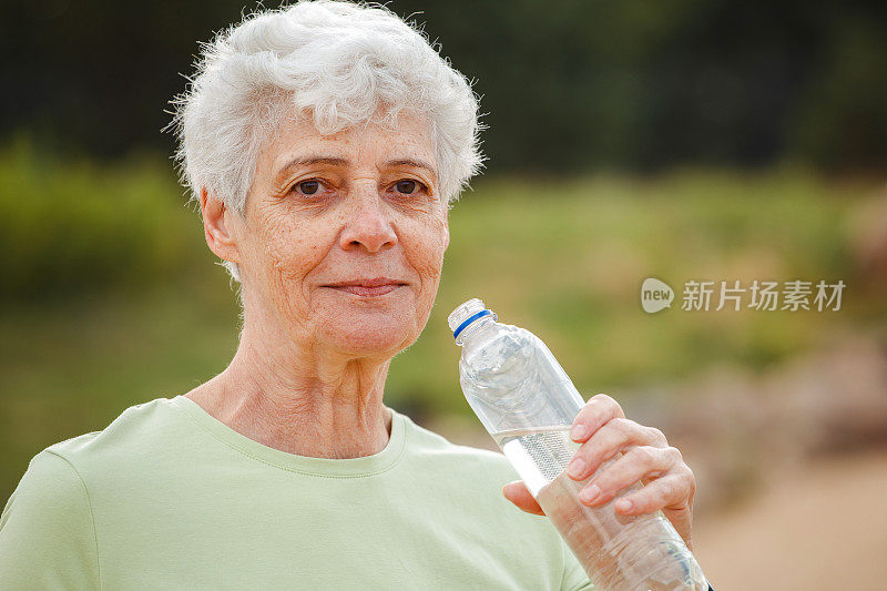 一位灰白短发的老妇人在运动后喝水，在公园里拍照