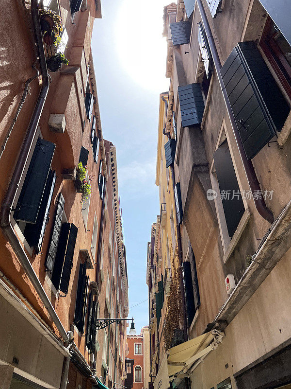 威尼斯滨水区狭窄的小巷里排列着建筑物的特写图片，打开百叶窗的多层窗户，阳光明媚的蓝天，从下面看