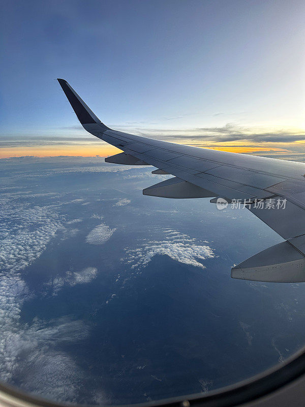 从飞机窗口看到的图像，飞机机翼在云层和大海之上，蓬松的白云在水面上，小翼和板条，橙色的日落，重点在前景