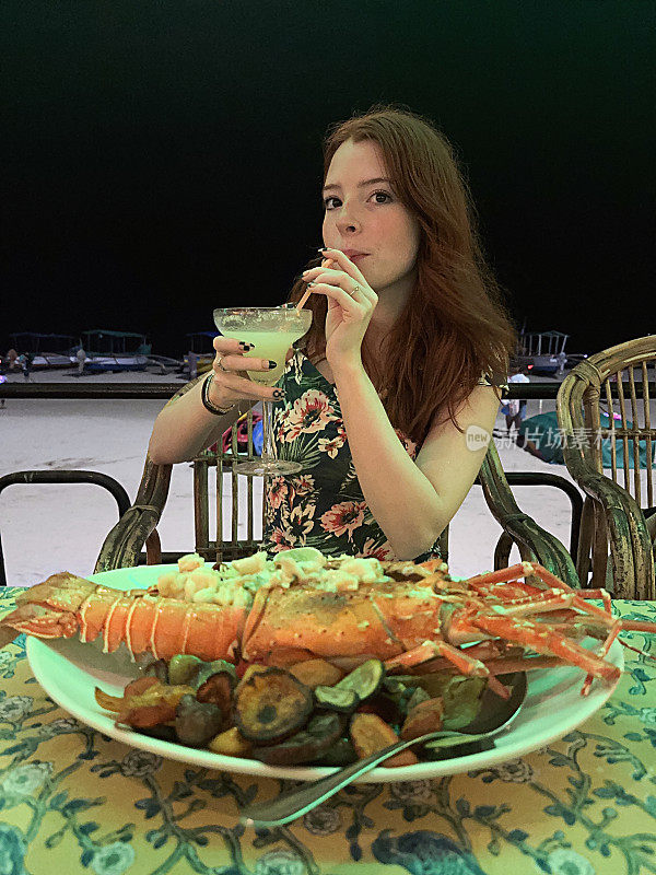 有吸引力的红头发的女人喝着玛格丽塔龙舌兰酒为基础的鸡尾酒与塑料吸管，玛格丽塔玻璃，户外穿着印花背心裙，新鲜出炉的热月龙虾，露天海滩餐厅在晚上，重点在前景