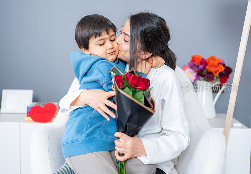 可爱的小男孩向妈妈祝贺，并送上一束玫瑰花。妈妈和男孩微笑着拥抱着。互相拥抱。母亲亲吻她的小儿子。母亲节快乐。