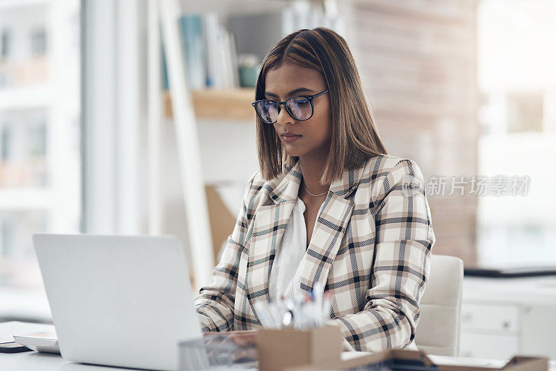 照片中，一位迷人的年轻女商人独自坐在办公室里，使用着她的笔记本电脑