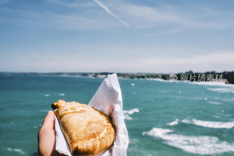 六月阳光明媚的纽基，一只手举着康沃尔馅饼在蓝色的海面上。