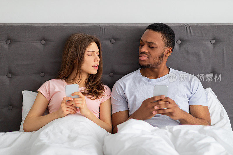 年轻的异族夫妇坐在床上，拿着智能手机，用怀疑的眼神看着对方