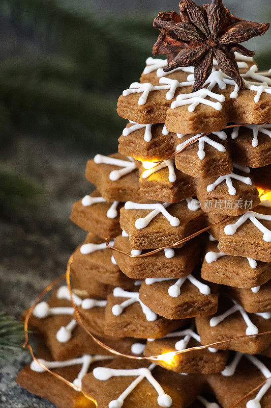 近距离拍摄的圣诞树是由姜饼雪花饼干堆叠而成，上面点缀着八角，装饰着白色的皇家糖衣和发光的仙女灯，灰色表面上是云杉针背景，聚焦前景
