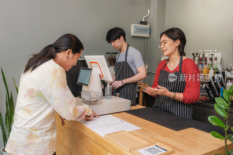 年轻的亚洲女性咖啡师或咖啡店老板在咖啡馆的吧台与她的客户讨论点饮料菜单