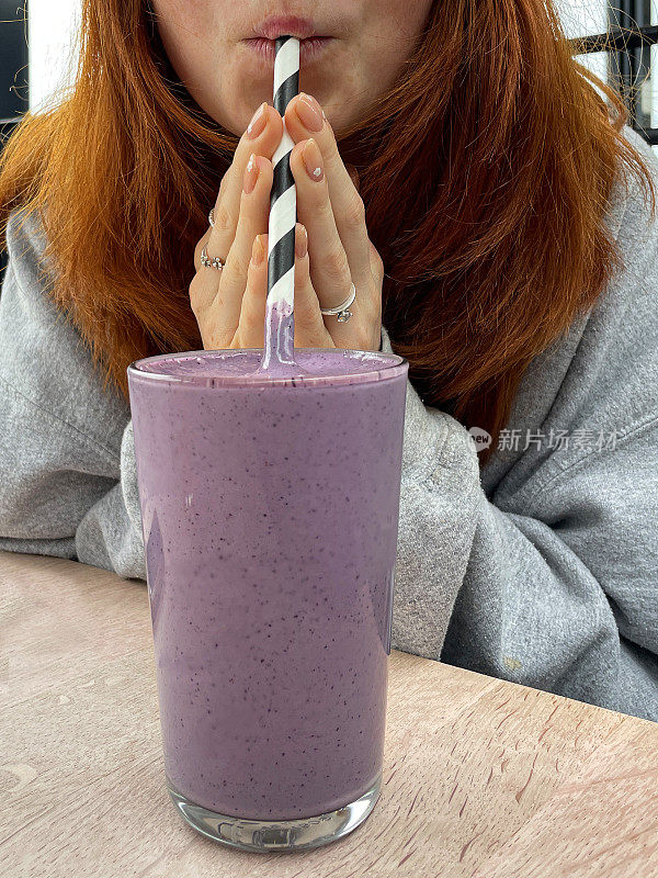 特写照片中，难以辨认的红发女子坐在餐厅的餐桌旁，喝着一杯蓝莓冰沙饮料，通过条纹状的纸吸管啜饮，聚焦于前景