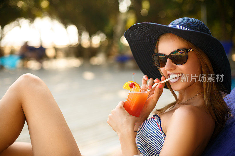 一个年轻女人的肖像与鸡尾酒杯在热带阳光下寒冷在游泳池附近的躺椅上，后面有棕榈树。假期的概念