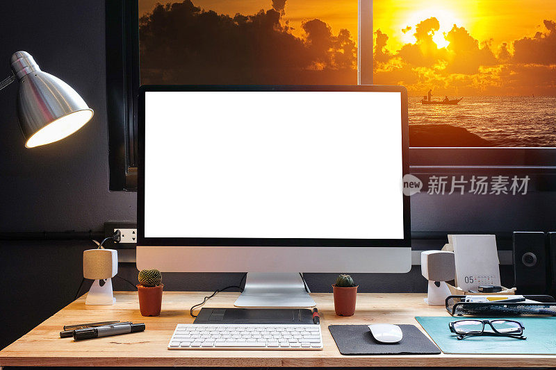 平面设计师的工作空间配备了一个手写板，一台电脑和白色的文本屏幕与美丽的日落从窗口的海滩