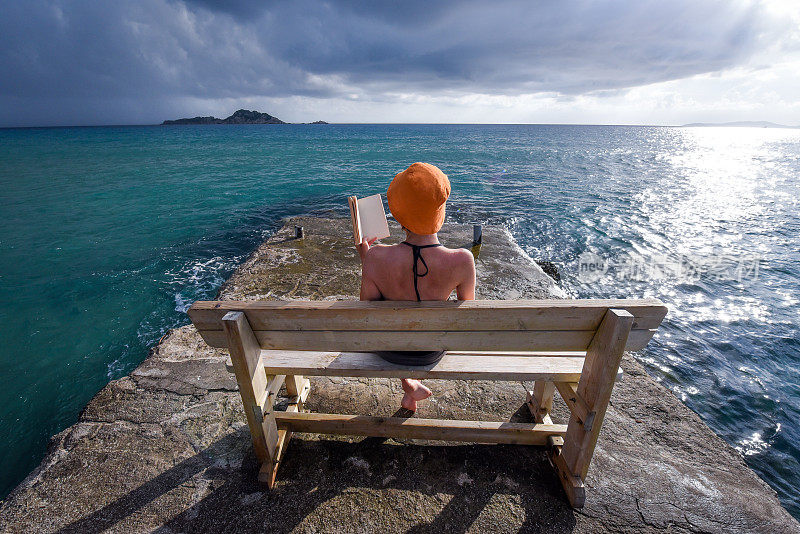 一名年轻女子在海边的长椅上看书，背景是引人注目的天空