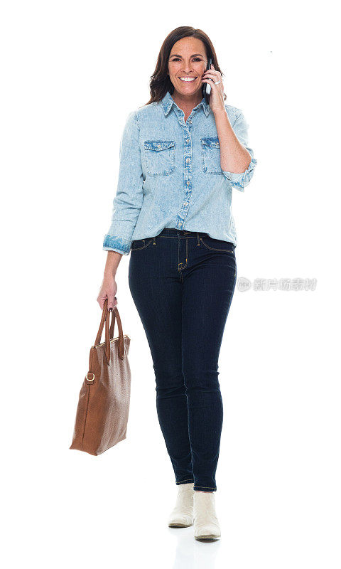 中年白人女性穿着牛仔裤走在白色背景前，拿着钱包，使用手机