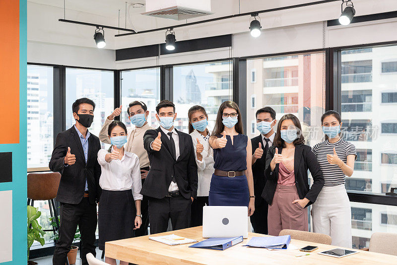 多民族商业团体自信地戴着口罩，在商业区新常态的办公室里竖起大拇指