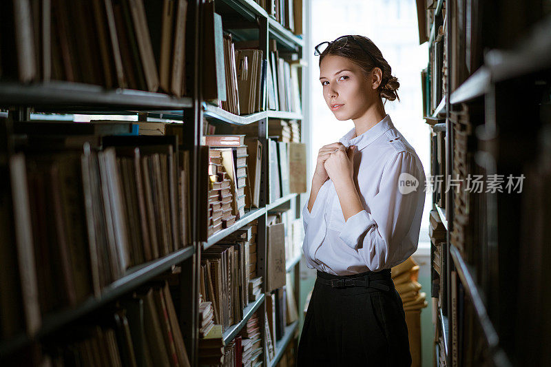 穿着白衬衫的漂亮女学生站在图书馆的两排中间，书架上放满了书。黑暗的照片