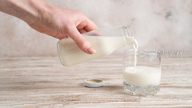 将牛奶倒入白色背景的玻璃杯中的玻璃瓶。手拿一瓶牛奶放在木桌上
