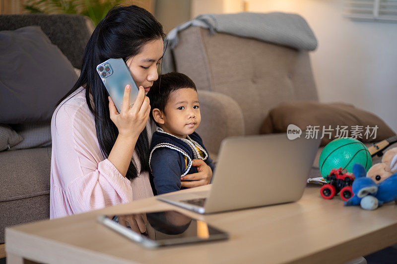 一个女人正在用笔记本电脑工作，一边打电话，一个男孩在他旁边玩。