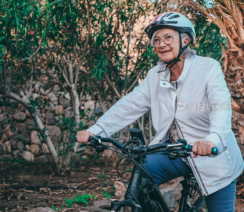 带着头盔的微笑老妇人骑着电动自行车在户外活动。老妇人在公园里骑自行车进行健康活动