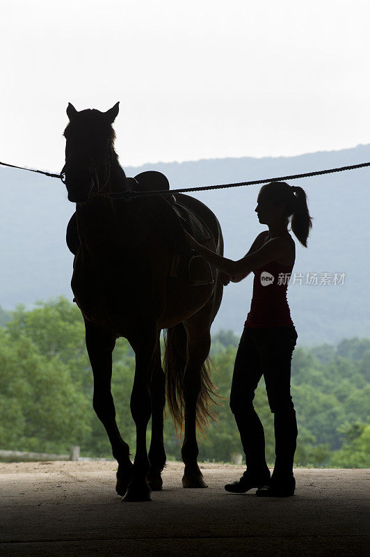 年轻女人和马的剪影在谷仓门，马鞍