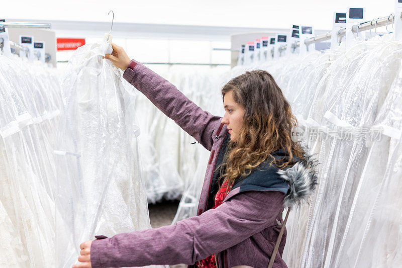 快乐的年轻女子拿着一件蕾丝婚纱在精品店折扣店，许多白色服装挂在衣架排上