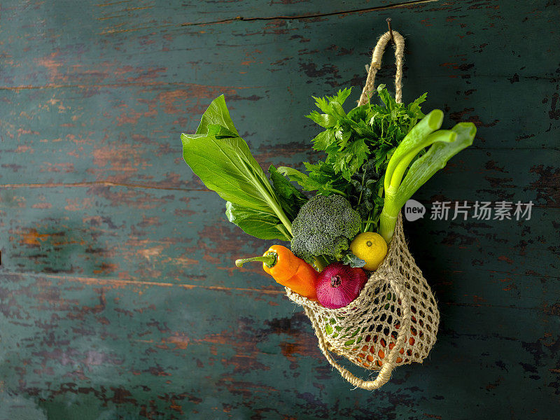 许多色彩鲜明的沙拉蔬菜在一个可重复使用的绳黄麻袋挂在旧风化的木头抽象颜色对比蓝绿色的背景墙。