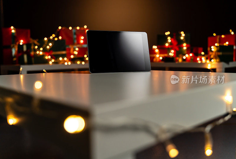 数码平板电脑在白色桌子前的散焦圣诞灯和圣诞礼物的背景，散焦