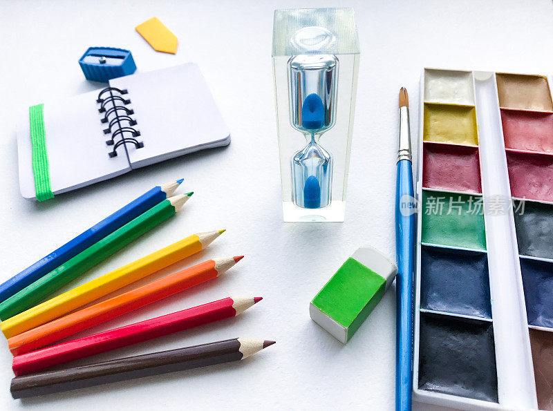 沙漏的特写和文具学校和创意，绘画和工艺品(水彩颜料，彩色铅笔，橡皮，卷笔刀)与白色纹理粗糙的水彩纸