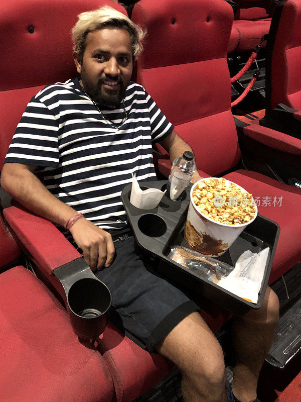 印度亚裔男子在电影院的座位上拿着一个托盘，里面装着零食、饮水机和爆米花