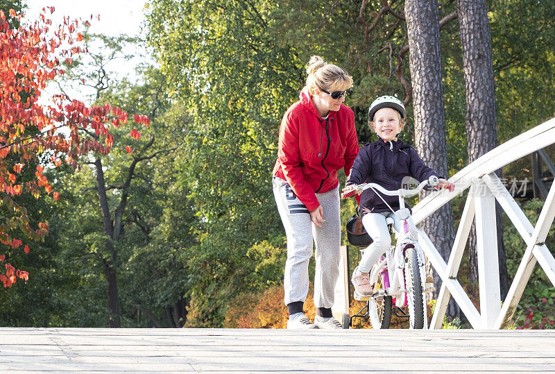 一个白人家庭在秋天公园的木制人行桥上散步。妈妈和女儿，一个骑自行车的孩子。