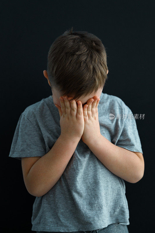 一个7岁的男孩站在黑色背景的灰色t恤上，完全是一个人，他用双手捂住脸，这个孩子正在经历情绪压力和哭泣。